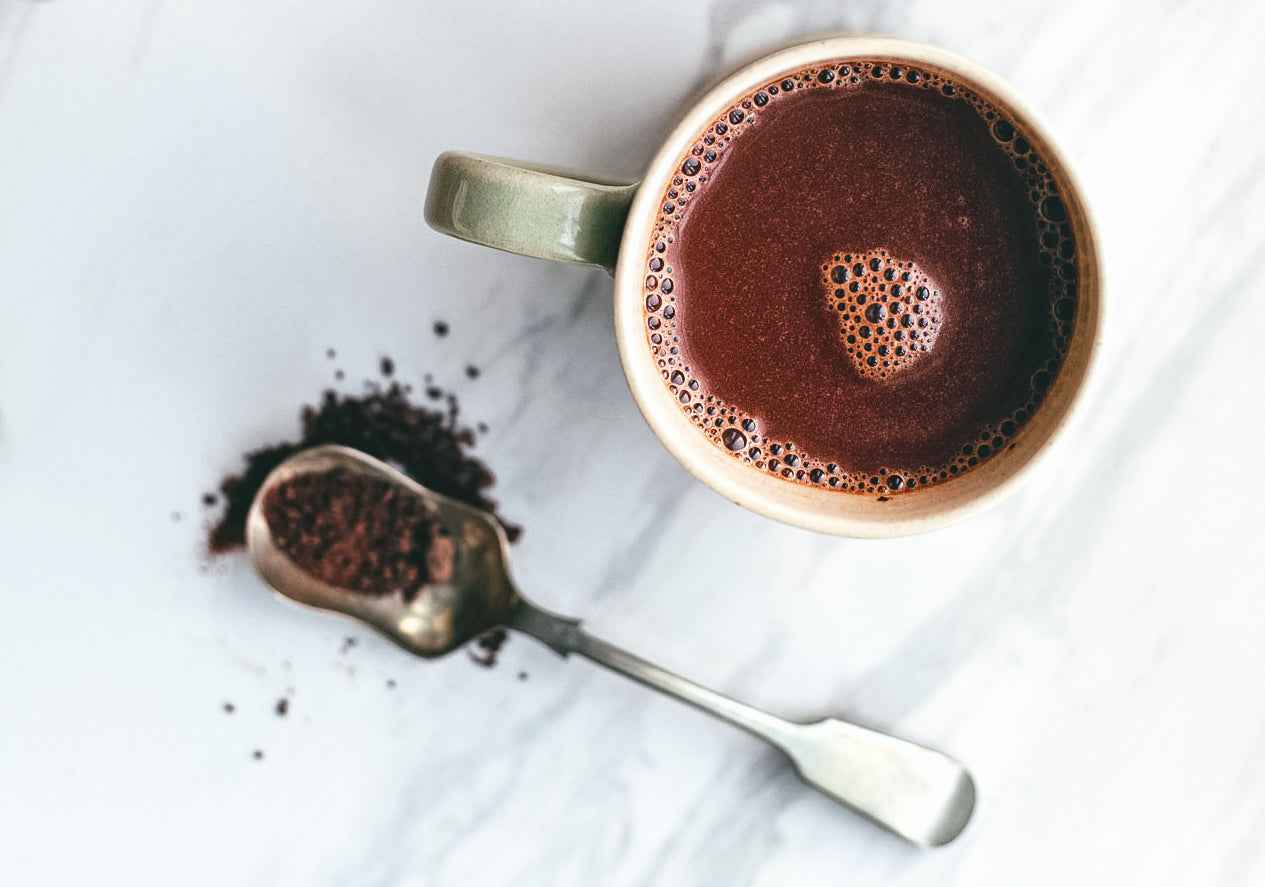 Scene depicting a cup from over head filled with warm Dominican Republic Zorzal Drinking Chocolate 70%, with a out of focus spoon containing chocolate granules strewn over a marble surface.