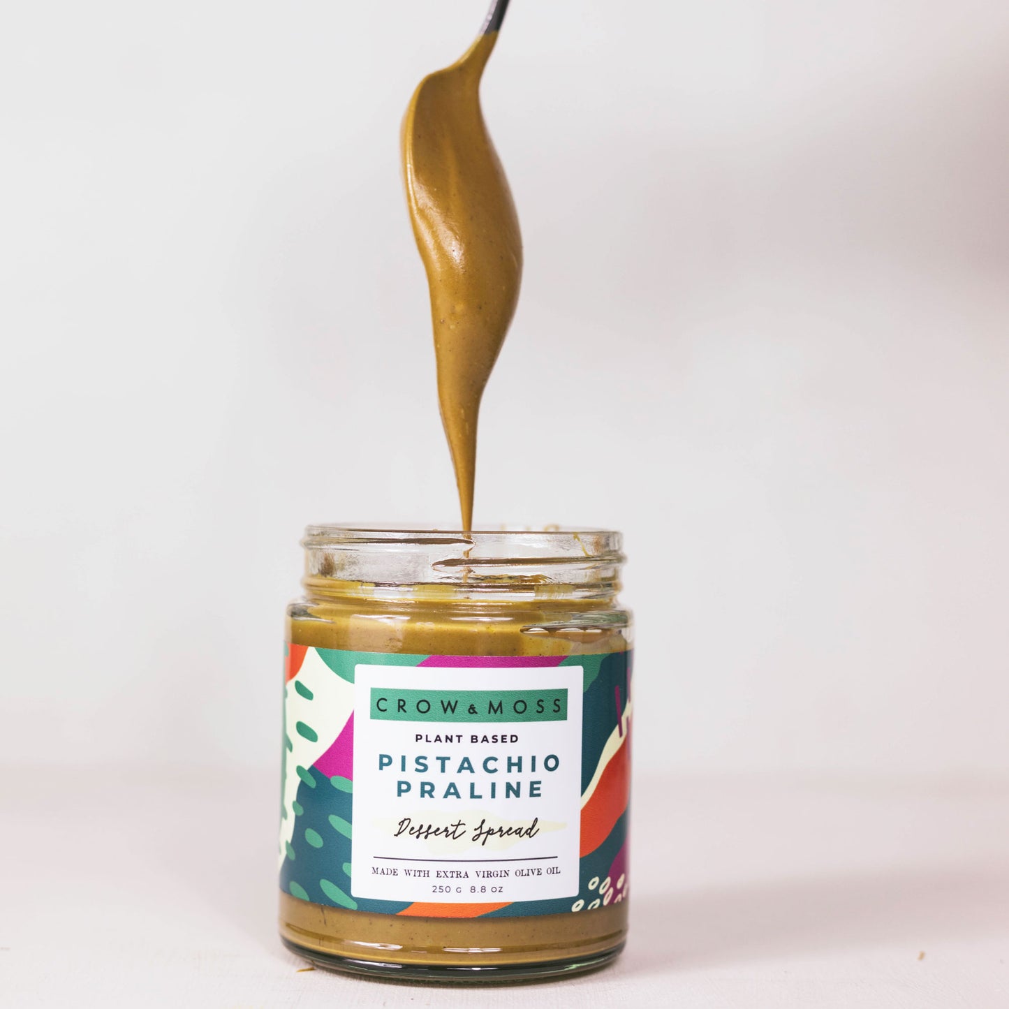 Jar of our Pistachio Praline Dessert Spread on a light green background with a spoon coming out of a jar allowing the paste to hang in the air momentarily.