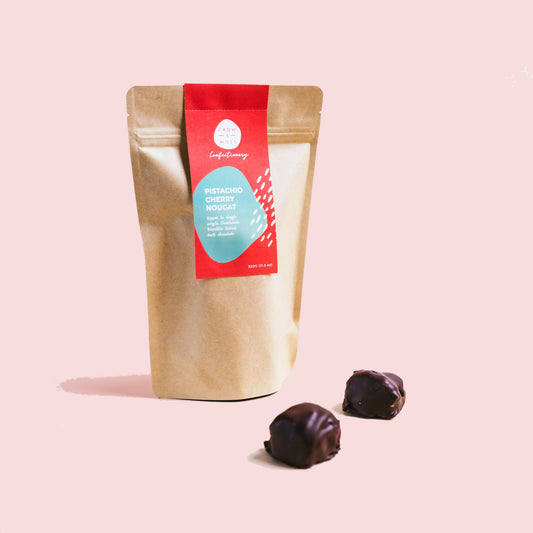 Image of a bag of our Chocolate Covered Pistachio Cherry Nougat with 2 Nougats in the foreground.